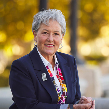 Della Gilleran is a middle-aged woman with short silver and gray hair. She is wearing a navy blue blazer, white-collared blouse, and a colorful scarf with a square pattern.