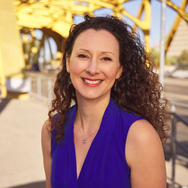 Isla Waite is a woman with long, curly brown hair. She is wearing a short-sleeved indigo top and a small necklace. In the background is a gold-colored bridge.