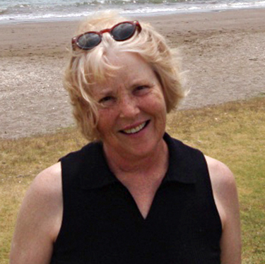 Jane Lorand standing on grass near a sandy beach. She has short blonde hair and a pair of sunglasses atop her head, wearing a short-sleeved black top. She is smiling at the camera.