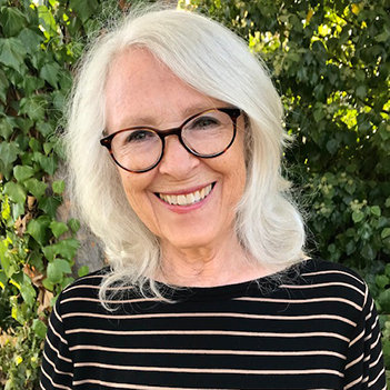 Karen Olson is a middle-aged woman with shoulder-length, wavy silver hair. She is standing in front of ivy foliage, is wearing black-rimmed glasses, a black top with horizontal peach stripes, and is smiling at the camera.