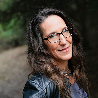 Kyra Auerbach is a middle-aged woman with long dark wavy hair. In the background is a dirt trail and trees. She is wearing dark-rimmed glasses, a dark top and dark jacket, with a beaded necklace.